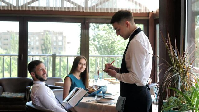 In A Restaurant, A Waiter Takes An Order From A Young Couple In An Elite Restaurant. Customer service. A waiter who gives recommendations in a restaurant.The customer reads the menu and makes an order