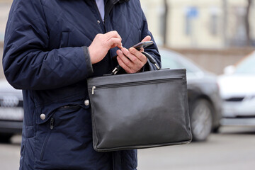 Man in coat standing with a leather briefcase with smartphone in hand on a city street. Concept of...