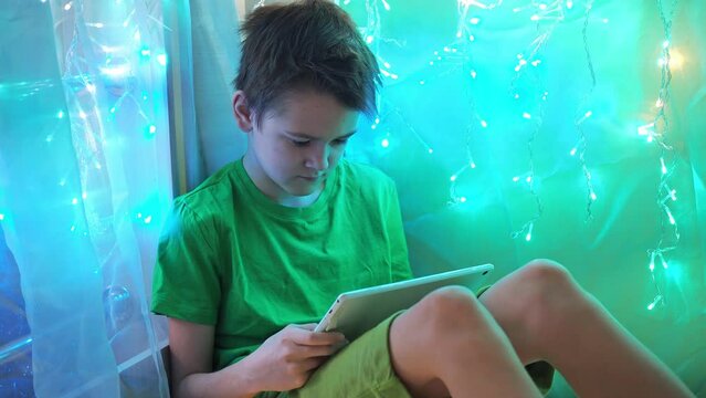 the boy looks at the tablet while sitting on a bed decorated with garlands. multicolored light of lights. a teenager in a green T-shirt and shorts