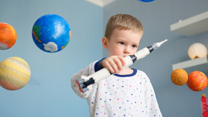 Little blond boy is playing with toy space rocket, flying in space among planets of solar system....