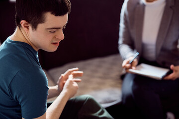 Shy down syndrome man during conversation with psychologist at home. - obrazy, fototapety, plakaty