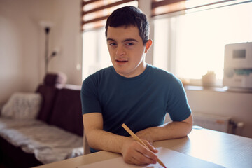 Obraz na płótnie Canvas Young down syndrome man writes on piece of paper at home and looking at camera.