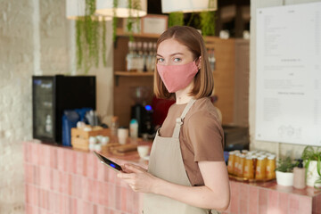 Horizontal medium portrait shot of young woman wearing apron and protective mask starting workday in cafe holding digital tablet looking at camera