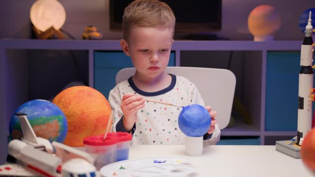 Blond boy paint planet solar system Pluto with colorful paint sitting home table in evening, planet solar system, spaceships and space shuttle from constructor around. Cosmonautics Day on April 12.