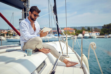 A young man watching a tablet content while sitting on a yacht and riding through the dock on the seaside. Summer, sea, vacation - obrazy, fototapety, plakaty