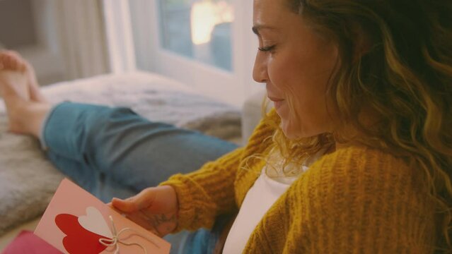 Young woman in cosy warm jumper lying on sofa at home opening and reading romantic valentine's day card - shot in slow motion