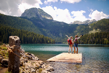 Hikers on wooden jetty by lake jumping in joy. Summer trip in nature. Lifestyle, love, togetherness, nature concept