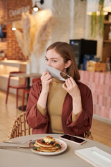 Vertical medium portrait of young Caucasian woman sitting at table taking off mask to eat meal in cafe