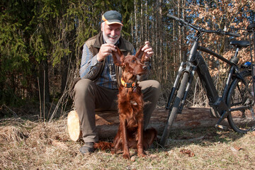 Dogs make people happy. Cyclist sits at the edge of the forest and has fun with his dog. He holds up his Irish Setter's floppy ears and laughs heartily.