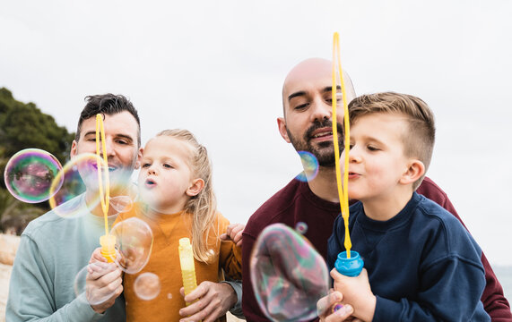 Gay Male Couple Playing With Son Children At The Beach With Bubble Soap Toy - LGBT Family Love Concept - Focus On Left Kid Face