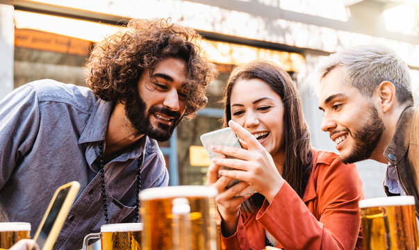 Young happy people having fun using mobile phone while drinking beer at brewery bar outdoor - Focus on center girl face