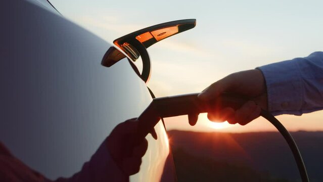 Unrecognizable businessman plugging electric car from charging station at sunset