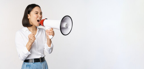Enthusiastic asian woman, girl activist shouting at protest, using megaphone, looking confident, talking in loudspeaker, protesting, standing over white background - obrazy, fototapety, plakaty