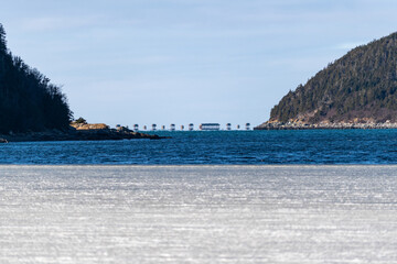 Obraz premium A wide view of frozen ice or slob in a cove. This ice has patches of snow with a glass surface. There are mountains or hills in the distance that are covered in green evergreen trees with blue sky.
