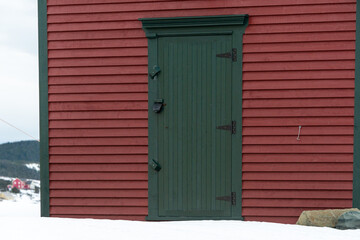 A winter's scene of a vintage dark green single wood door with black hinges and handle. The wall is covered in a red narrow cape cod clapboard siding. The edge or trim of the building is green.