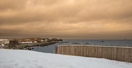 A breakwater made of wood logs along the edge of the ocean with a bank of white snow in the foreground. The sky is golden with heavy clouds. There's a landmass in the distance with a rocky edge.  
