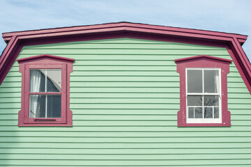 A vibrant lime green exterior wall of narrow horizontal cape cod clapboard on a house. There are two single four pane glass casement windows with bright pink decorative trim with white lace curtains
