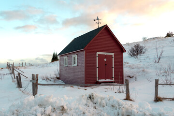 A small red wooden barn or shed in the middle of a snow covered fenced in field. There's a small hill behind the building. There's a metal weather vane on top of the red building with white trim.  - obrazy, fototapety, plakaty