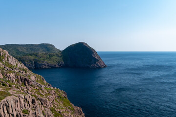 A scenic oceanview of hills, rocky cliffs, and treed covered mountains in Newfoundland. The view from the hiking trail is of a calm blue sea. The horizon is a light color with a blue sky. 