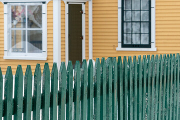 Obraz premium A bright and cheerful yellow country house with wood cape cod clapboard, white trim, and multiple windows. There's a green wooden picket fence in the front of the building enclosing the garden.