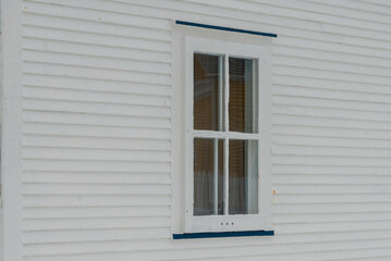 A small single casement window in a white narrow wood beveled clapboard siding exterior wall of a vintage house. The glass window has six panes with green decorative trim on the outside edge. 