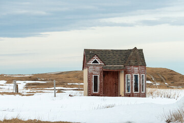 A small vacant house with a brown wooden exterior, single level, turret, peaked roof, and long casement windows. The quaint building is in a meadow covered in white snow.There's a blue sky with clouds - Powered by Adobe
