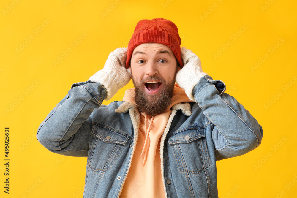 Wall mural Surprised young bearded man in warm gloves and hat on yellow background