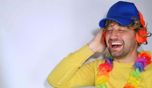 Image of handsome man in casual and party outfit, wearing a blue cap, listening music in orange headphones, smiling pleased, happy standing over white background