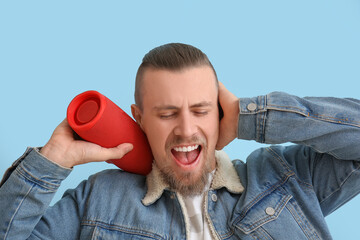 Emotional man holding wireless portable speaker and closing ears on blue background