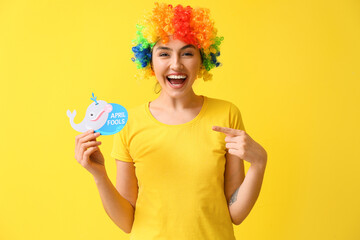 Funny young woman in colorful wig and with paper fish on yellow background. April fools' day celebration