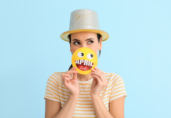 Young woman holding paper smile with text APRIL FOOLS on blue background