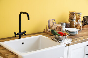 Fototapeta na wymiar White sink, colander with vegetables and kitchen utensils on counters near yellow wall