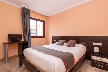 Hotel room with double with wooden headboard and brown cushions with red decorations and dark marble floor, desk with flat tv and brown aluminum window with gray curtains