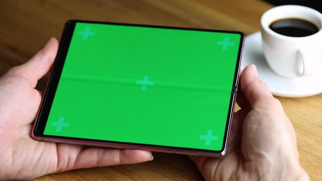 New folding Smartphone Samsung Galaxy Z Fold 2 with green screen, cup of coffee lying on wooden table, woman's hand double tapping on. Flat lay top view, mock up.