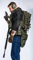 airsoft player in full growth. a man in an outfit, in headphones, a bulletproof vest, with a rifle and a pistol on a white background. stands sideways, in profile