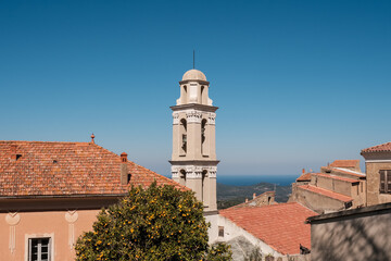 Fototapeta premium The bell tower and terracotta roof tiles in the village of Costa in the Balagne region of Corsica with an orange tree in the foreground and Mediterranean sea in the distance