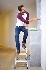 Getting into the corners with the perfect brush. Photo of an attractive young man painting a wall indoors while sitting on a ladder.