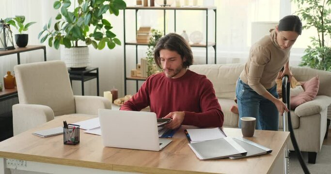 Woman asking working husband to get up while vacuum cleaning at home