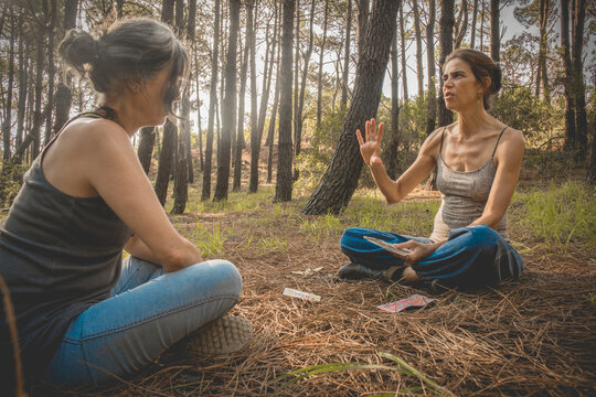 A female tarot-card reader is taking about the future to her girlfriend sitting on the ground in a spiritual moment into the woods during springtime.