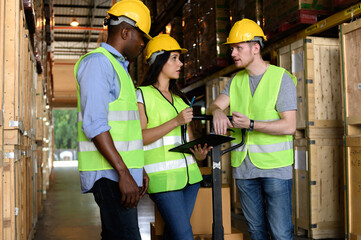 Diversity group of warehouse worker emplyee wearing hard hat and safety tools pushes pallet truck and a stack of boxes in a warehouse discussing and talking about work process schedule.