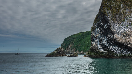 Naklejka premium A picturesque rocky island in the Pacific Ocean. Seabirds nest on the bare stones of steep slopes. Cloudy. A lonely boat in the distance. Kamchatka. Starichkov Island