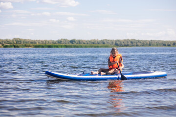 Female child sup boarding on her own on calm lake with oar in hands looking at water ripples in vest life jacket. Active holidays. Inculcation of love for sports from childhood.