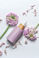 Pastel purple cosmetic bottle with pipette and beautiful flowers and petals on white background. Natural cosmetic and skin care routine. Beauty product. Face serum. Top view