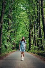 Fototapeta premium Young 20-24 year old slim brunette in a light blue summer dress dances on the road to the rhythm of the music. Candid portrait of a real ordinary person enjoying movement and dancing. Knowing oneself