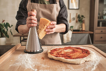 Close-up of unrecognizable woman in apron standing at table with wooden board and raw pizza and grating cheese on grater