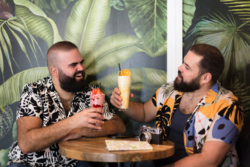 Two gay men enjoying their vacation together. They are happy toasting with a cocktail. Summer and travel as a couple. - obrazy, fototapety, plakaty