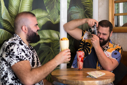 Young Gay Couple Taking Pictures While Drinking A Cocktail In A Tropical Bar. Tourism, Summer And Travel Concept.