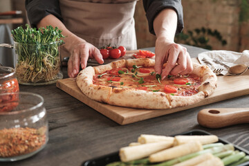 Close-up of unrecognizable woman in apron standing at table with jars of spices and decorating hot pizza with herbs