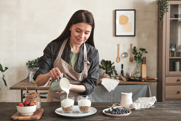 Content attractive young woman in apron standing at kitchen counter and pouring yogurt into mugs...