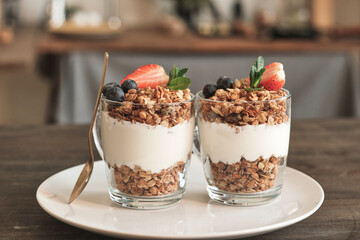 Close-up of two transparent mugs of granola with muesli and summer berries on white plate on dining...